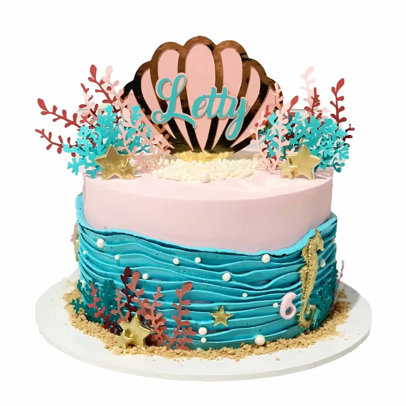 Pink Under the Sea Paradise Cake - Pink iced cake with blue piped waves, adorned with a custom shell cake topper, coral, seaweed, and pearls, a captivating choice for an undersea-themed celebration.