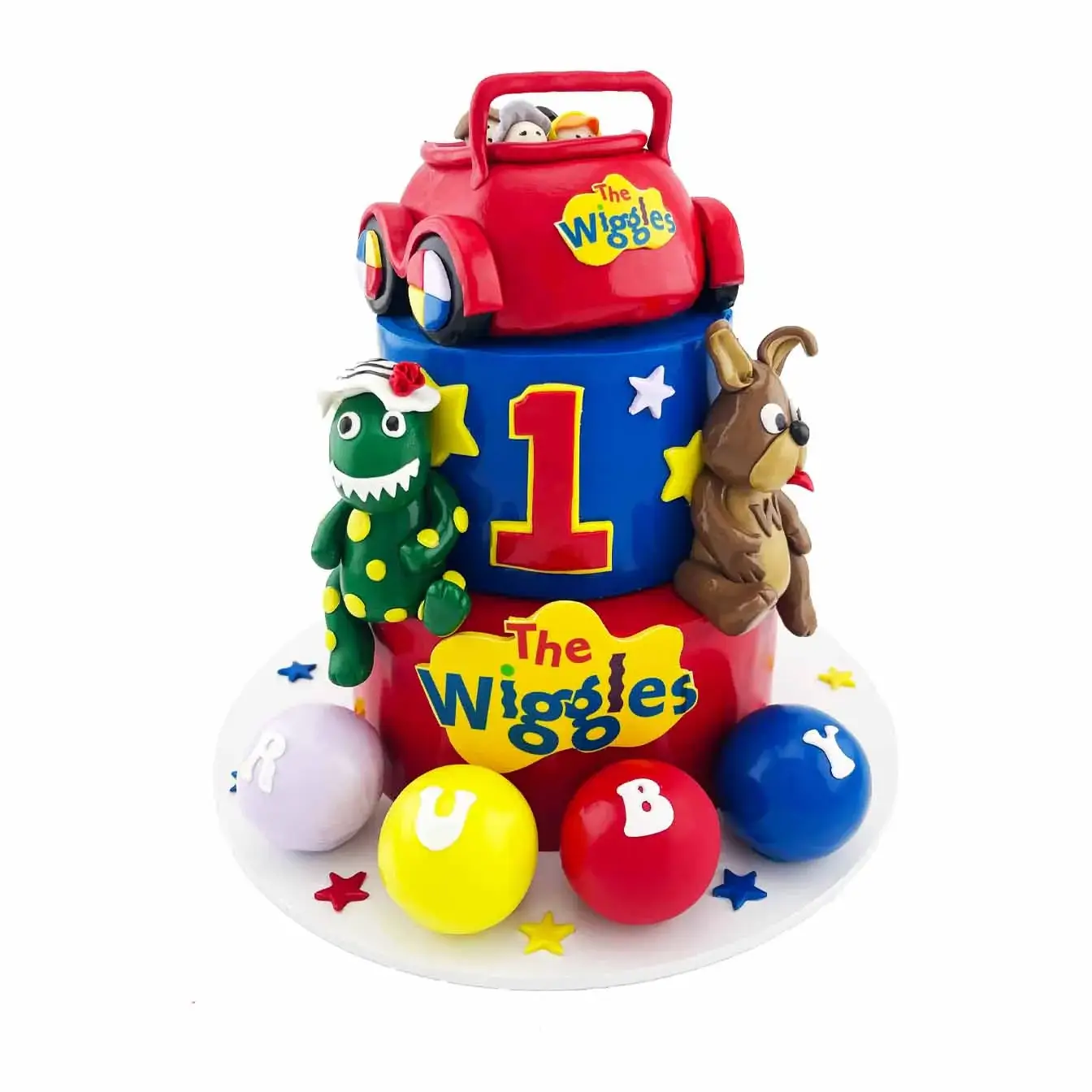 Wiggles Big Red Car Celebration Cake - A two-tier blue and red iced cake with a moulded Big Red Car, Wiggles logo, cutout stars, Dorothy the Dinosaur, Wags the Dog, colored balls, and personalized name, a delightful centerpiece for a Wiggles-themed celebration.